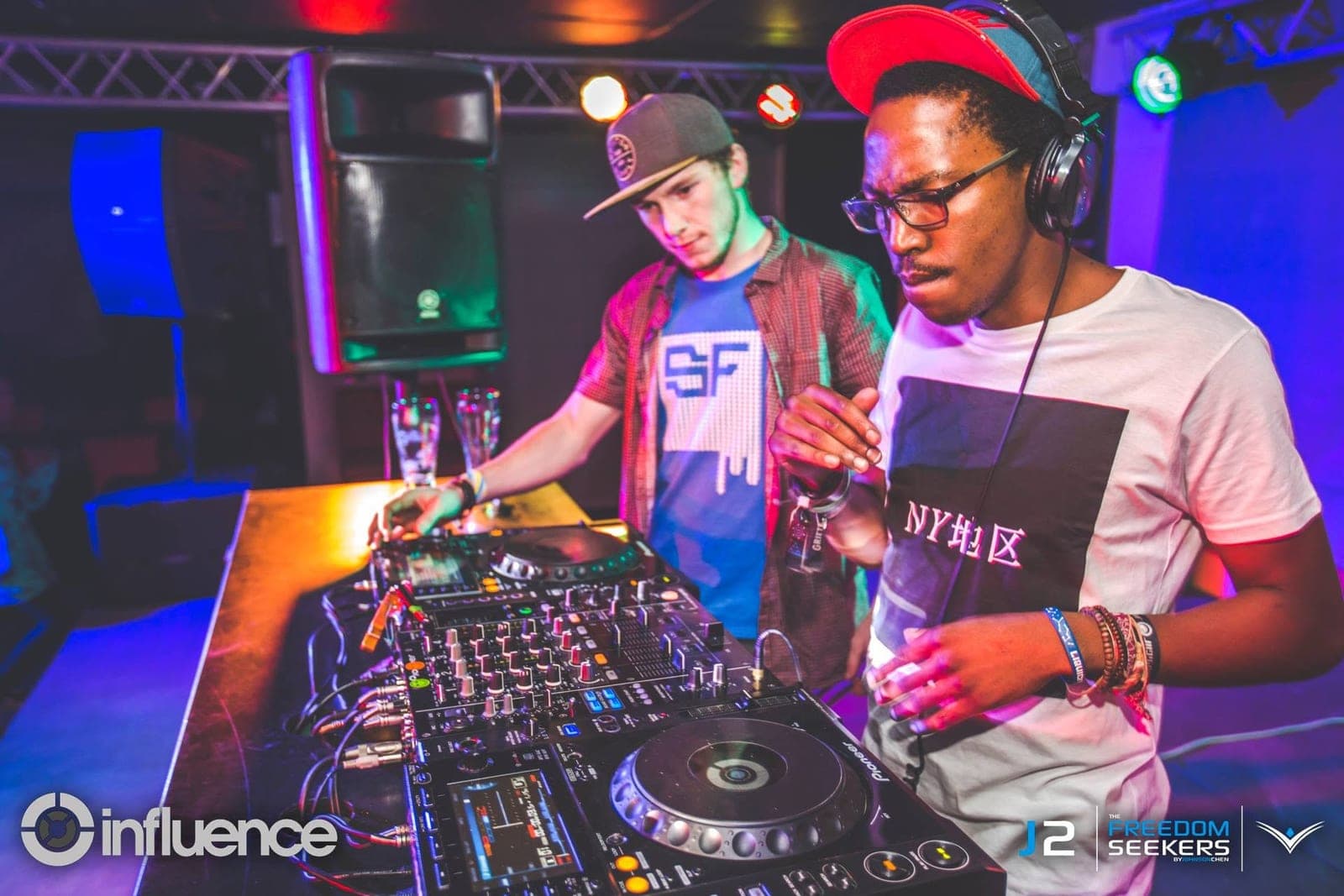 Two DJs at the decks under blue and pink light, one in a red cap and glasses focused on the mixer.