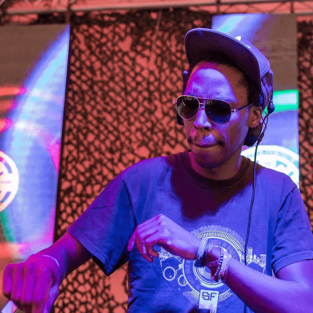 A close portrait of the DJ in sunglasses and a cap, lit in purple and red stage light.