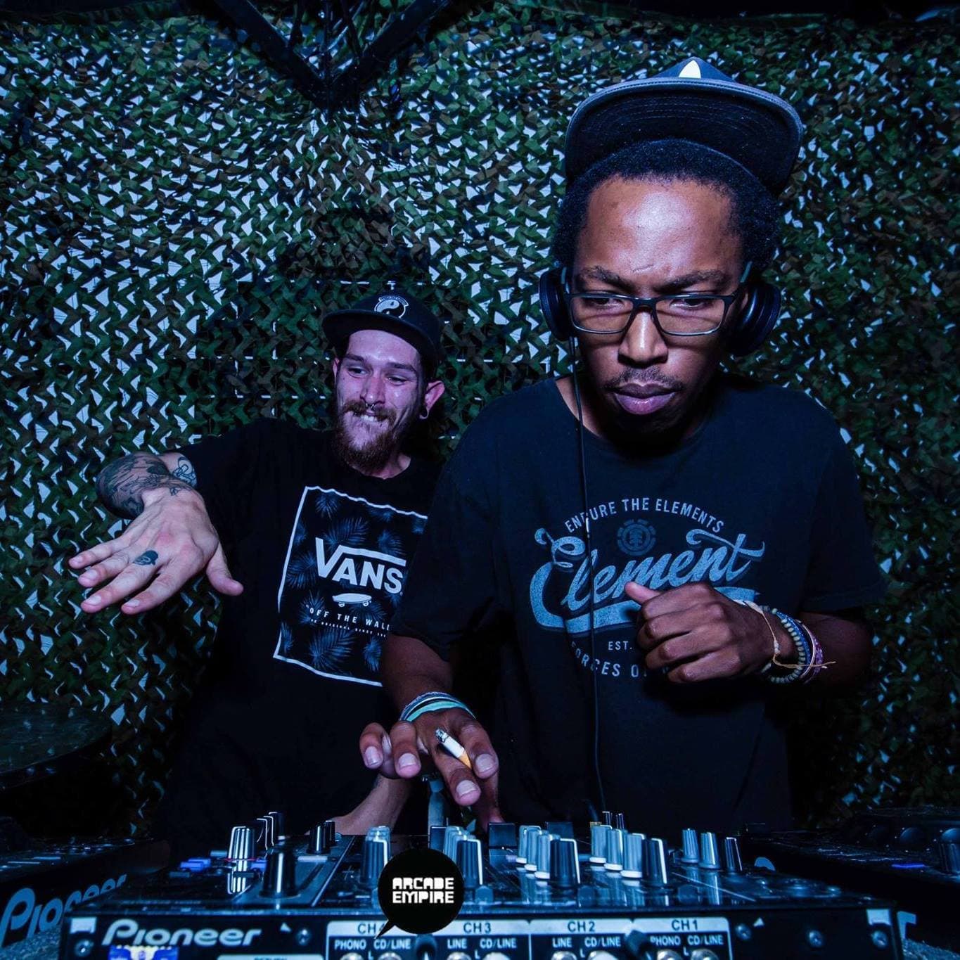 The DJ at the decks in a camouflage-netted booth, with another attendee beside him.