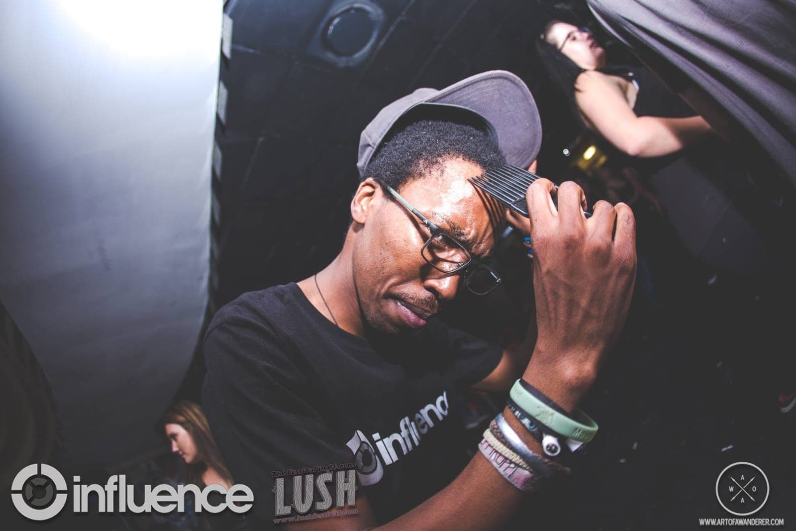 A DJ in an Influence t-shirt and grey cap squints and runs an afro comb through his hair behind the decks.
