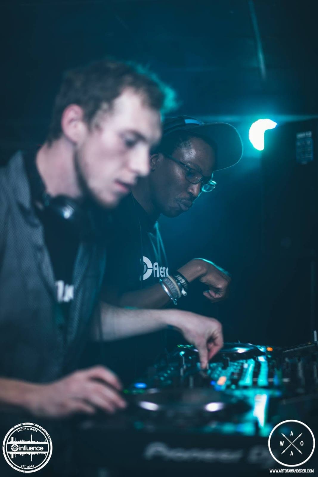 Two DJs share the decks under teal light; the one in the foreground is out of focus and the one behind wears glasses and a cap.