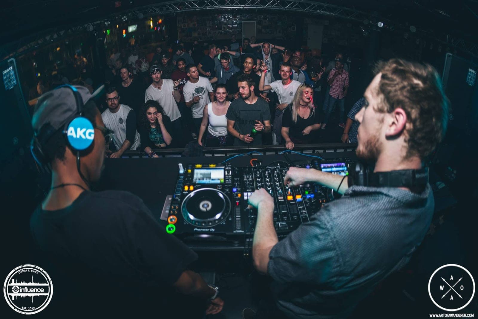 Shot over the DJs' shoulders, two DJs play to a smiling crowd pressed against the booth barrier.