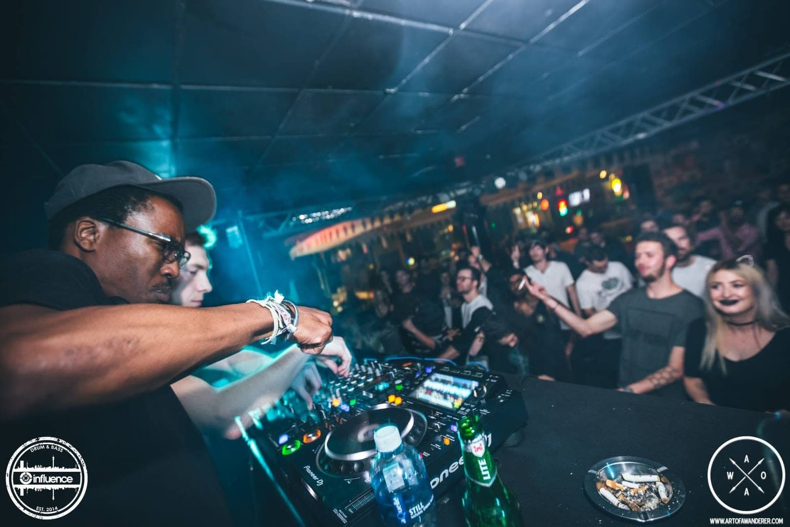 A DJ in glasses reaches across the mixer through club smoke while two DJs play to a crowd.