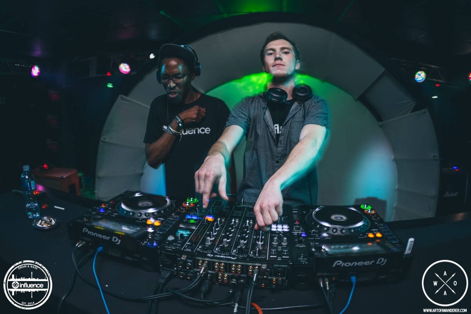 Two DJs pose at the Pioneer decks in front of a white dome, one in an Influence t-shirt, one in a grey shirt.