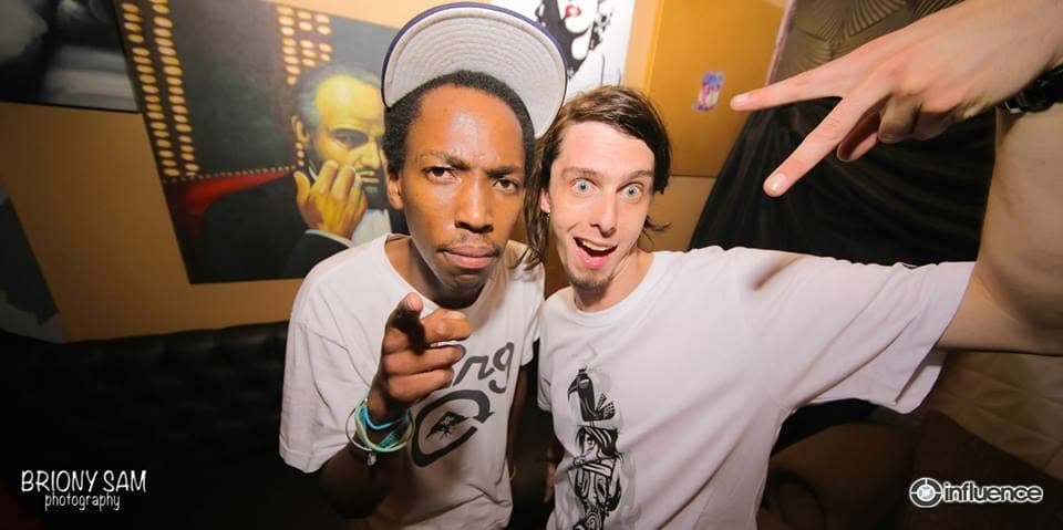 Two friends in white t-shirts pull faces at the camera, one pointing, in a room with a painted portrait behind them.