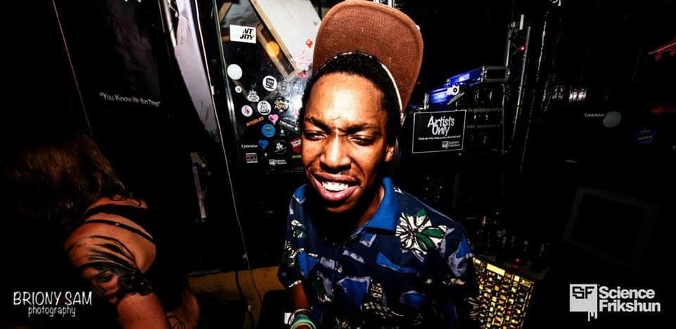 A man in a blue floral shirt and brown cap grimaces and grins backstage next to an "Artists Only" sign.