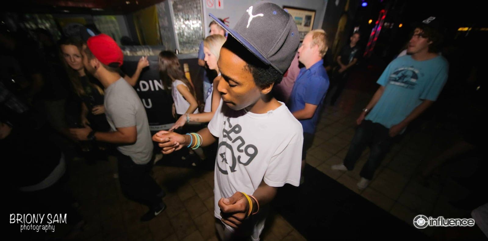 A man in a Puma cap and white t-shirt dances with his head down on a club floor among the crowd.