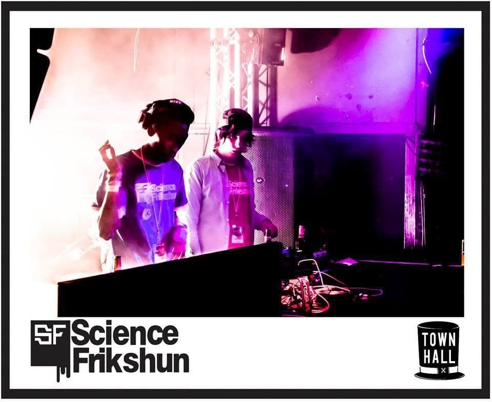 Two DJs in Science Frikshun shirts stand behind a booth lit in pink and purple haze.