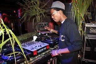 A DJ in a grey cap and glasses works a laptop-and-CDJ setup among tall plants.
