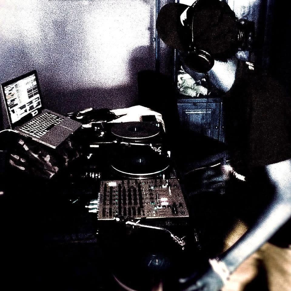 A heavily processed, grainy shot of a DJ in a cap and headphones beside turntables, a mixer and a laptop.