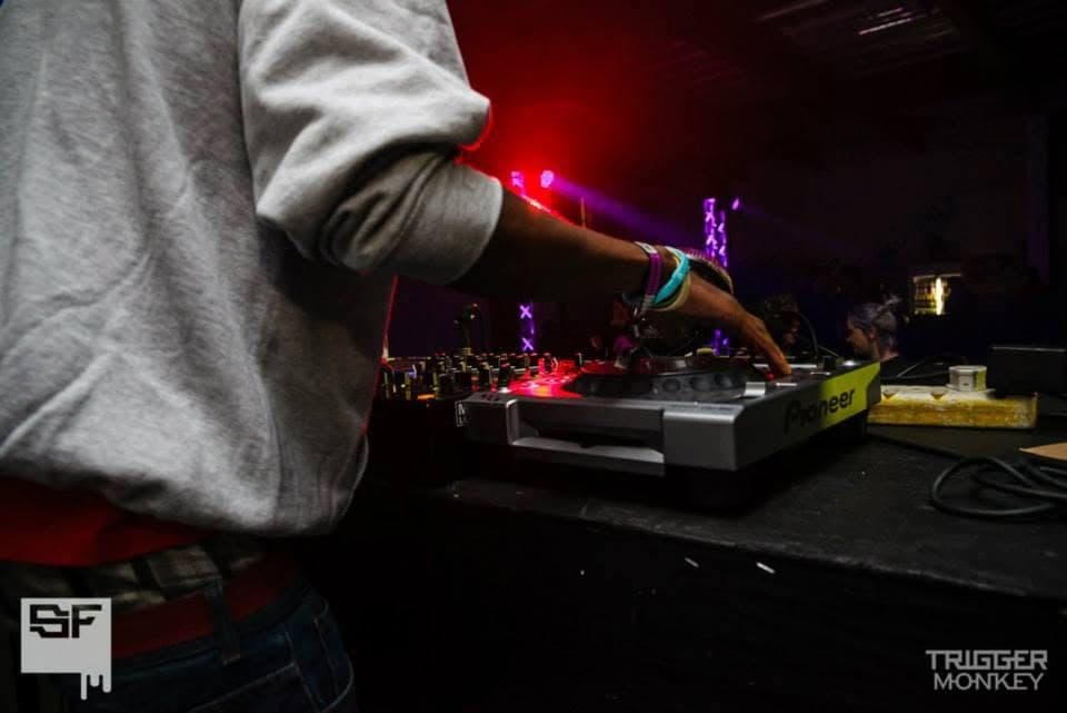A DJ in a grey hoodie is cropped to torso and forearm as he works a Pioneer CDJ under red club light.