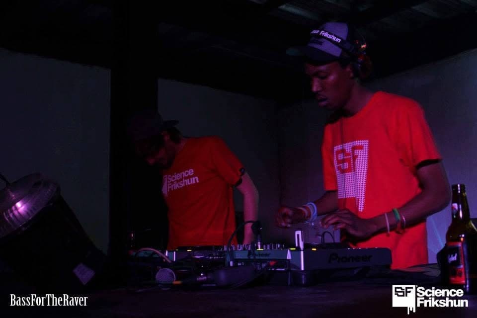 Two DJs in red Science Frikshun t-shirts behind a Pioneer mixer in a dim room.