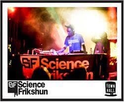 DJ playing behind a Science Frikshun-branded booth in smoky orange light.