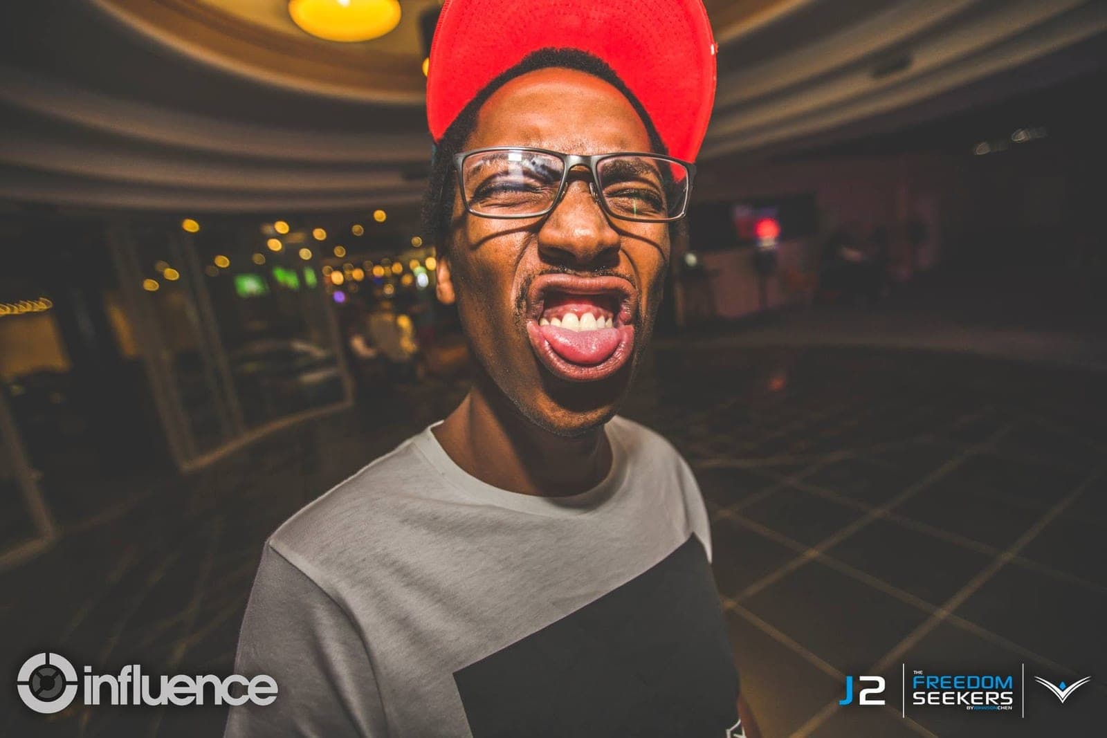 Close wide-angle portrait of a man in a red cap and glasses pulling a face, in a dim lounge.