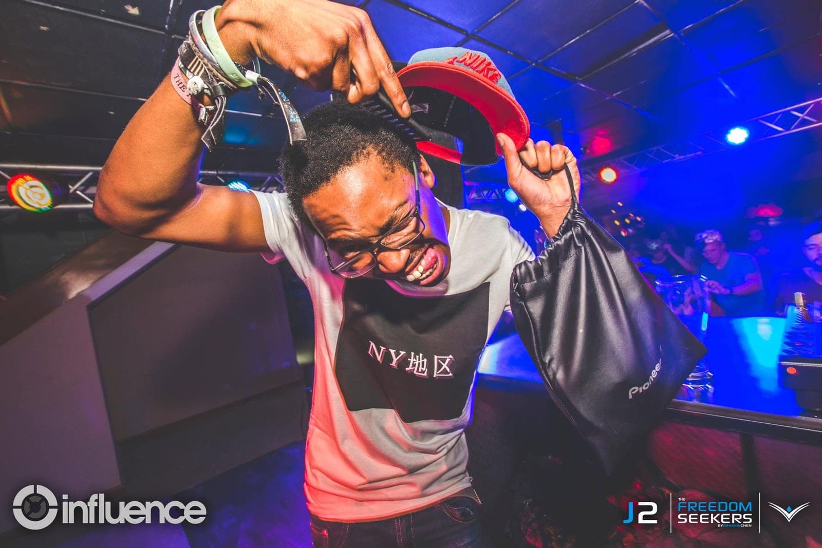 Man in an "NY" tee combing his hair while holding a red cap and a Pioneer bag near the DJ booth.