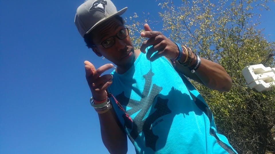 Man in a teal tank top, glasses and a grey cap pointing both hands at the camera against a blue sky.