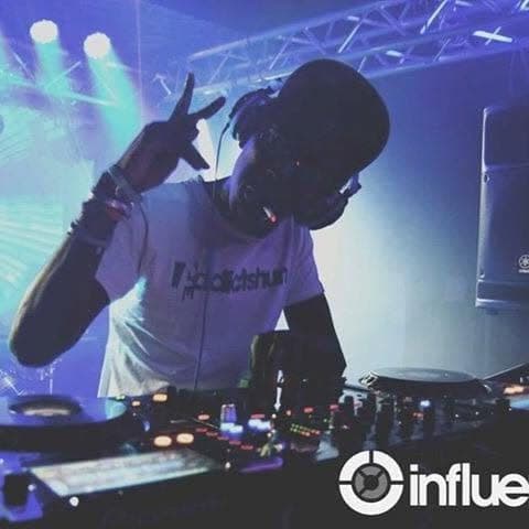 DJ in an Addictshun t-shirt flashing a peace sign over the decks under blue stage lights.
