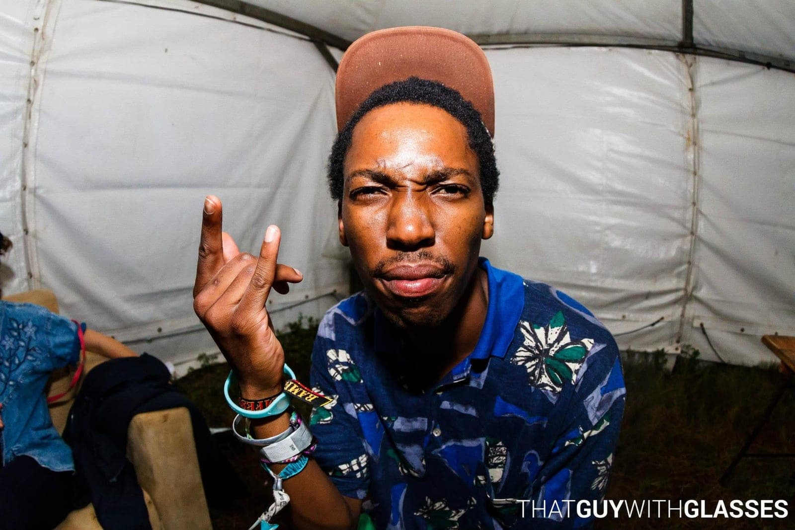 Man in a brown cap and blue patterned polo pulling a face and holding up a hand inside a festival tent.