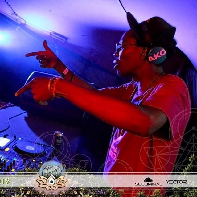 DJ in a red t-shirt and AKG headphones pointing both hands forward, lit in red and purple, with a lotus emblem below.