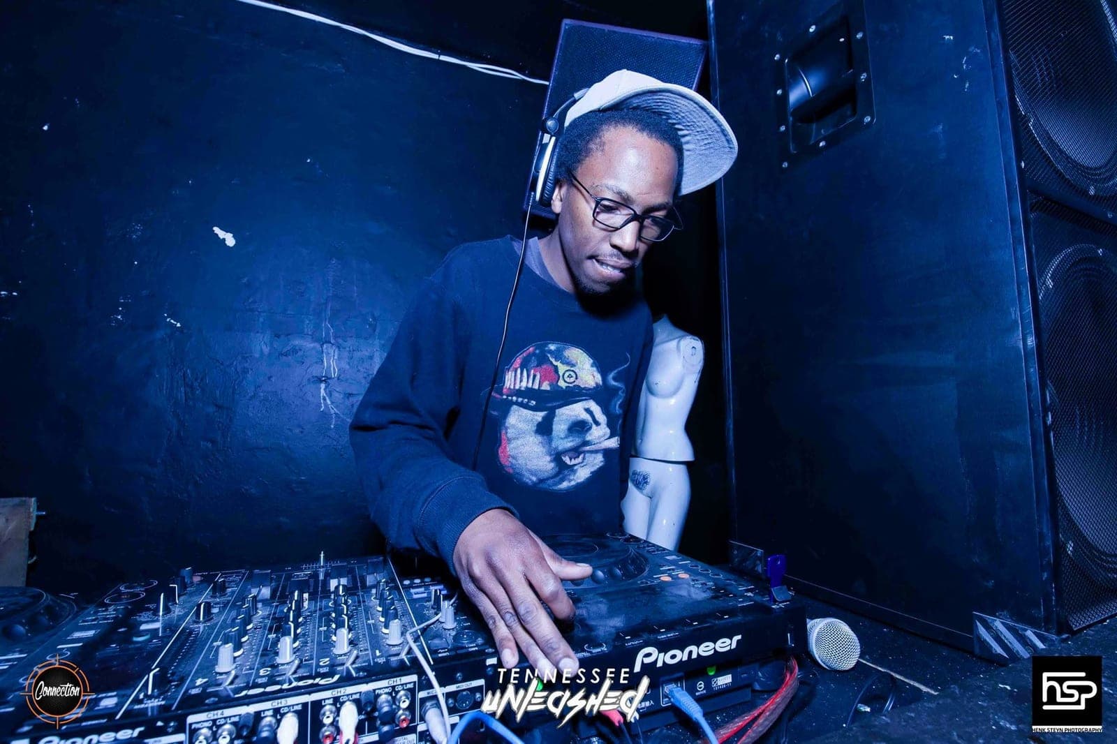 The DJ in glasses and a grey cap mixing on Pioneer decks in a dark, blue-lit booth beside stacked speakers.