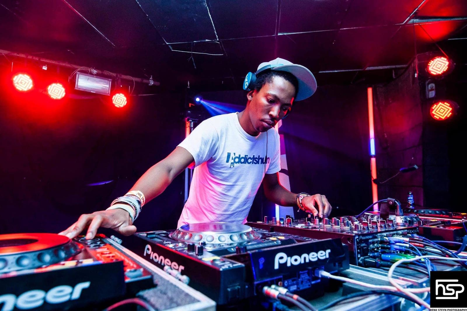 A DJ in an Addictshun t-shirt and pale cap leaning over Pioneer decks under red and purple stage lights.