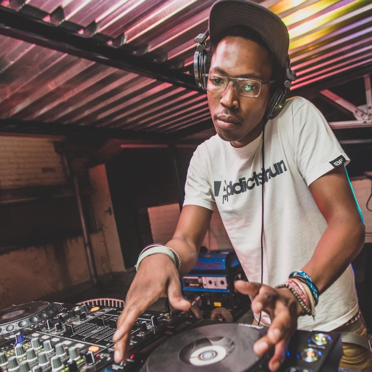 The DJ adjusting the mixer under a corrugated tin roof lit in pink and gold, headphones around his neck.