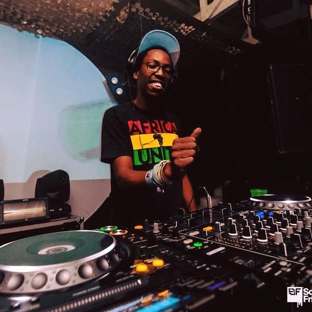 The DJ smiling and giving a thumbs up behind the decks, wearing an "Africa United" t-shirt.