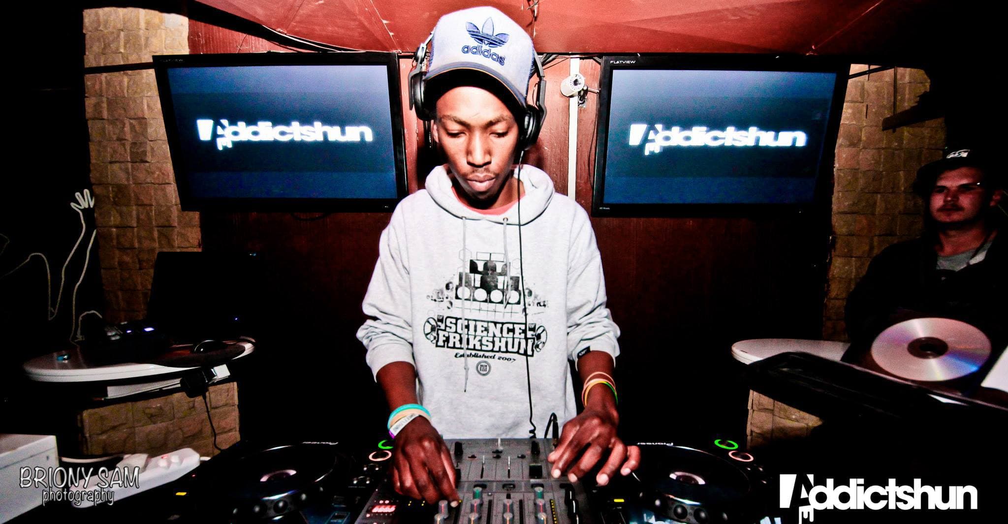 The DJ between two screens displaying event branding, wearing a grey hoodie.