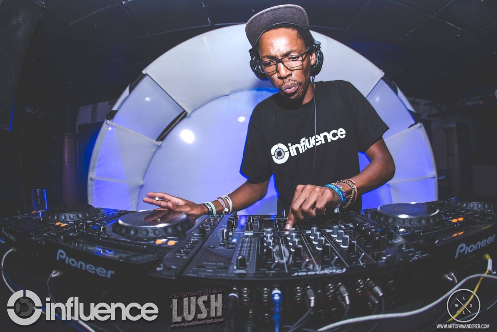 A DJ in glasses, a grey cap and an Influence t-shirt mixing in front of a white dome stage.
