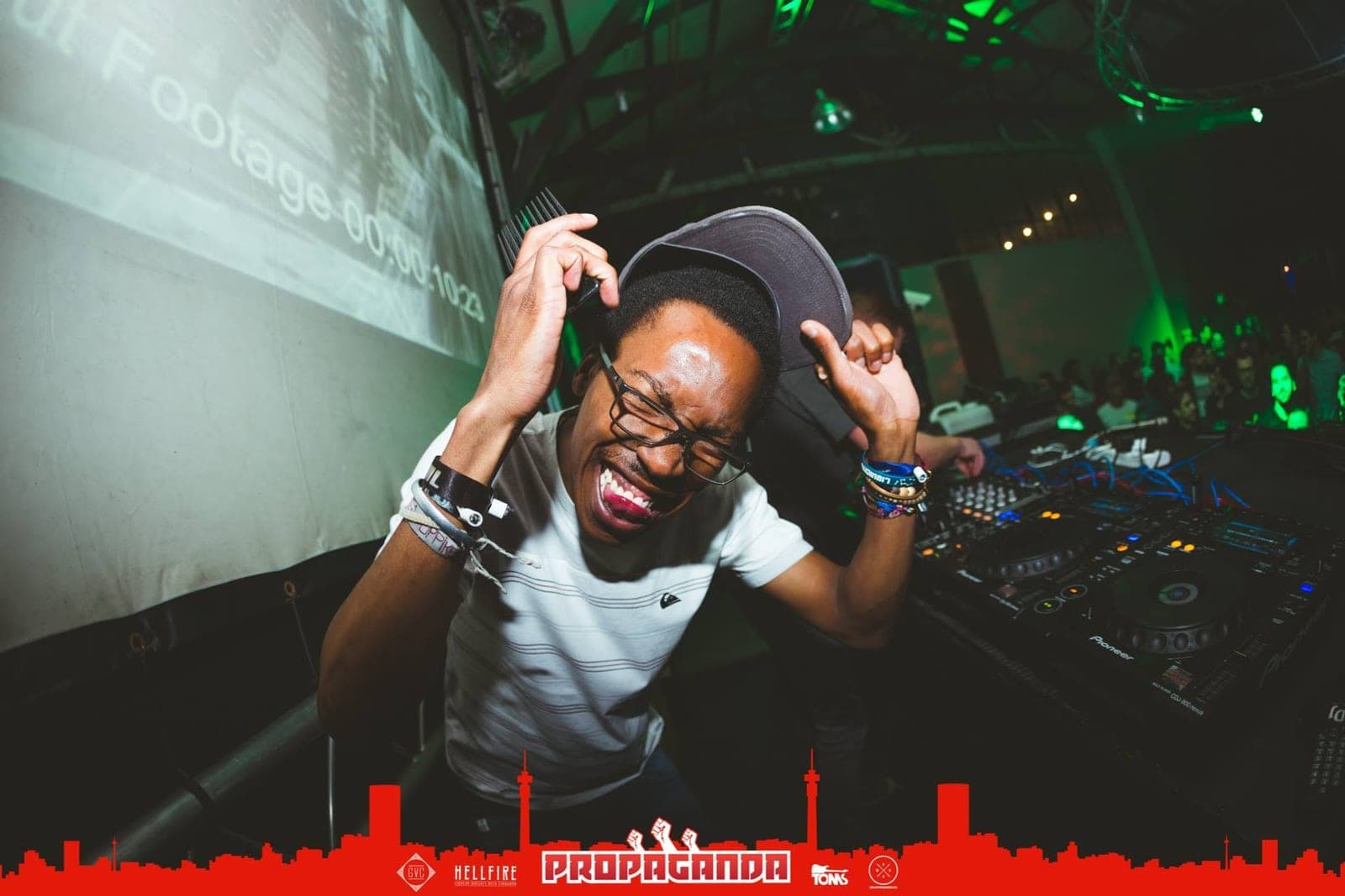 A DJ grins with his eyes shut, holding an afro comb and tilting his cap next to the decks.