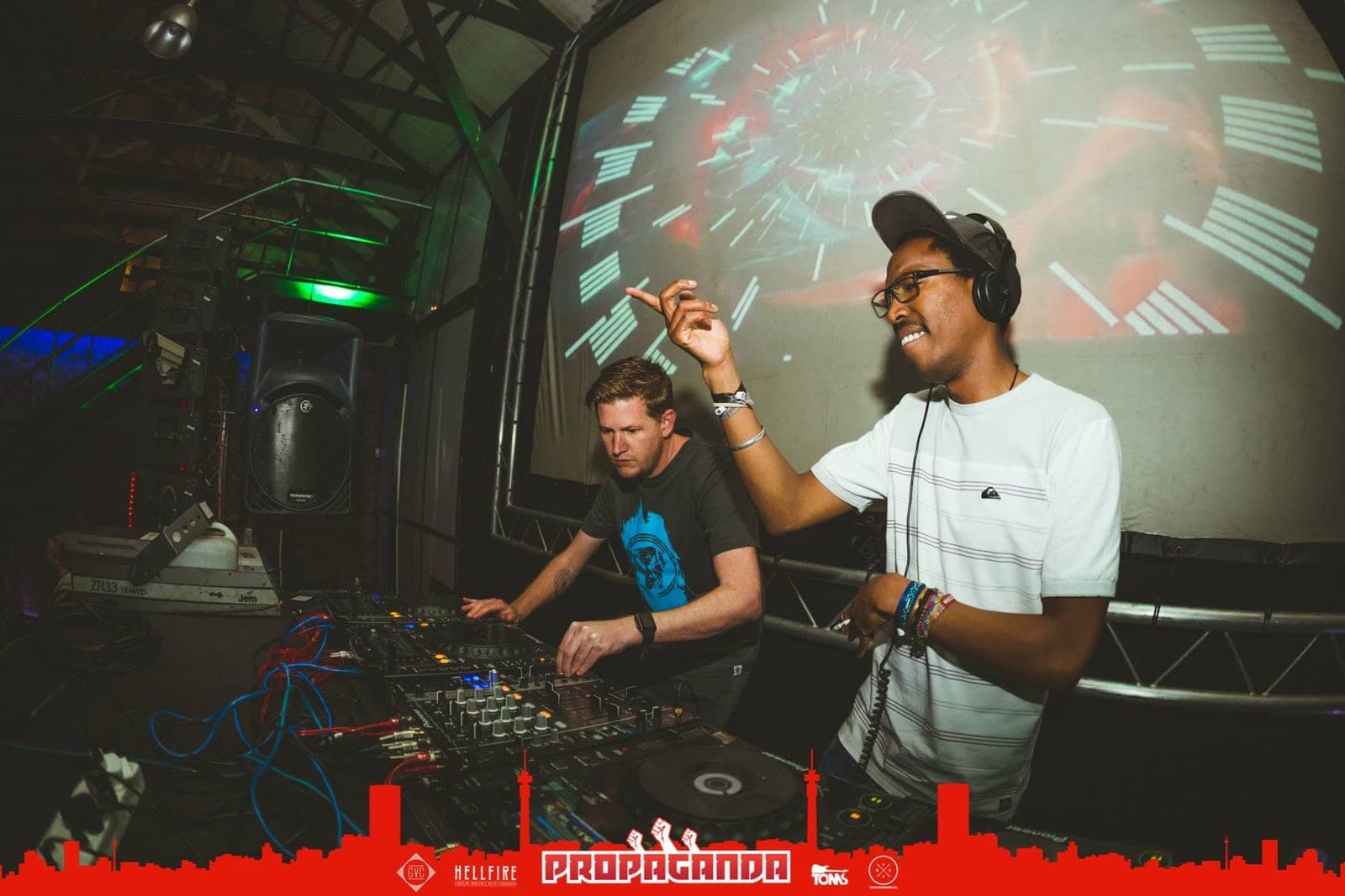 A smiling DJ in headphones points upward beside a second DJ in a dark t-shirt, with a video wall behind.