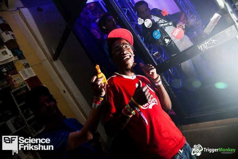 A grinning man in a red football shirt and red cap holds up a yellow rubber chicken and gives thumbs up.