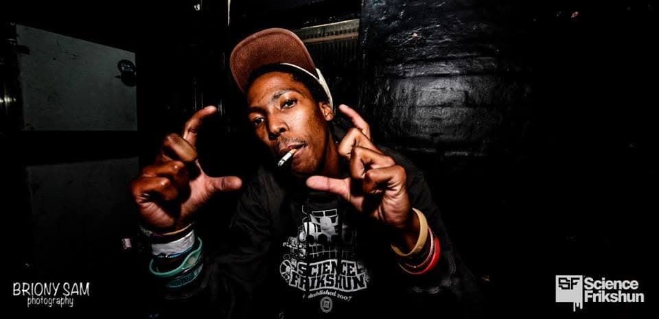 A man in a Science Frikshun shirt and brown cap holds a cigarette in his mouth and frames the camera with his hands.