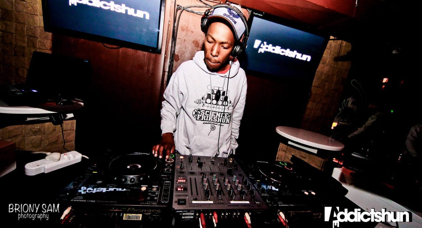 A DJ in an Adidas cap and Science Frikshun hoodie looks down at Pioneer CDJs and a mixer beneath two Addictshun screens.
