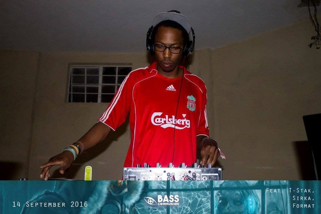 DJ in a red Liverpool shirt, glasses and headphones mixing in a bare room.