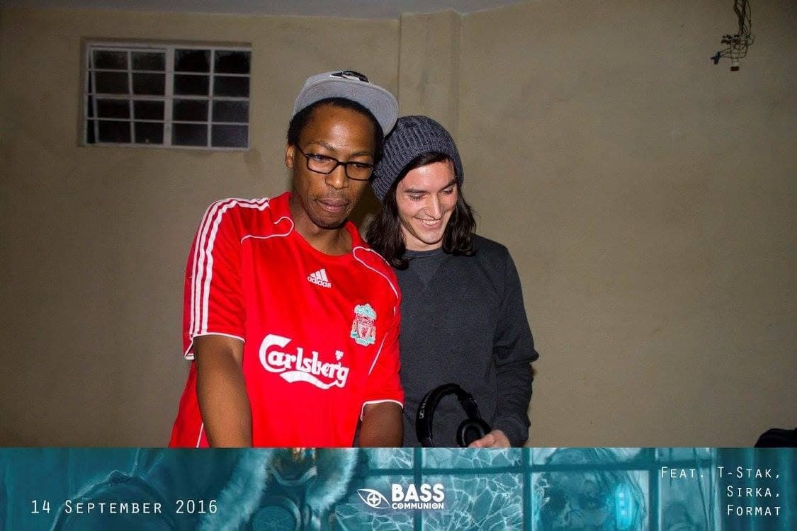 Two DJs, one in a red Liverpool shirt and one in a beanie, laughing together at the decks.