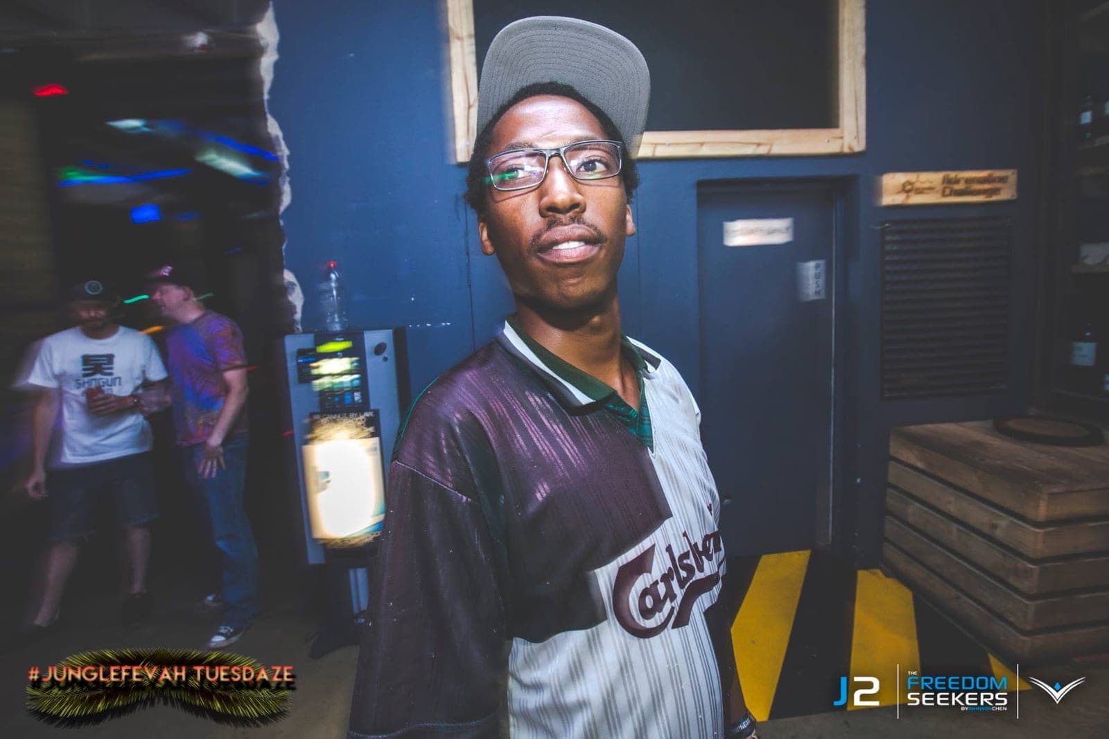 Man in glasses, a grey cap and a Carlsberg football shirt looking sideways in a club corridor.