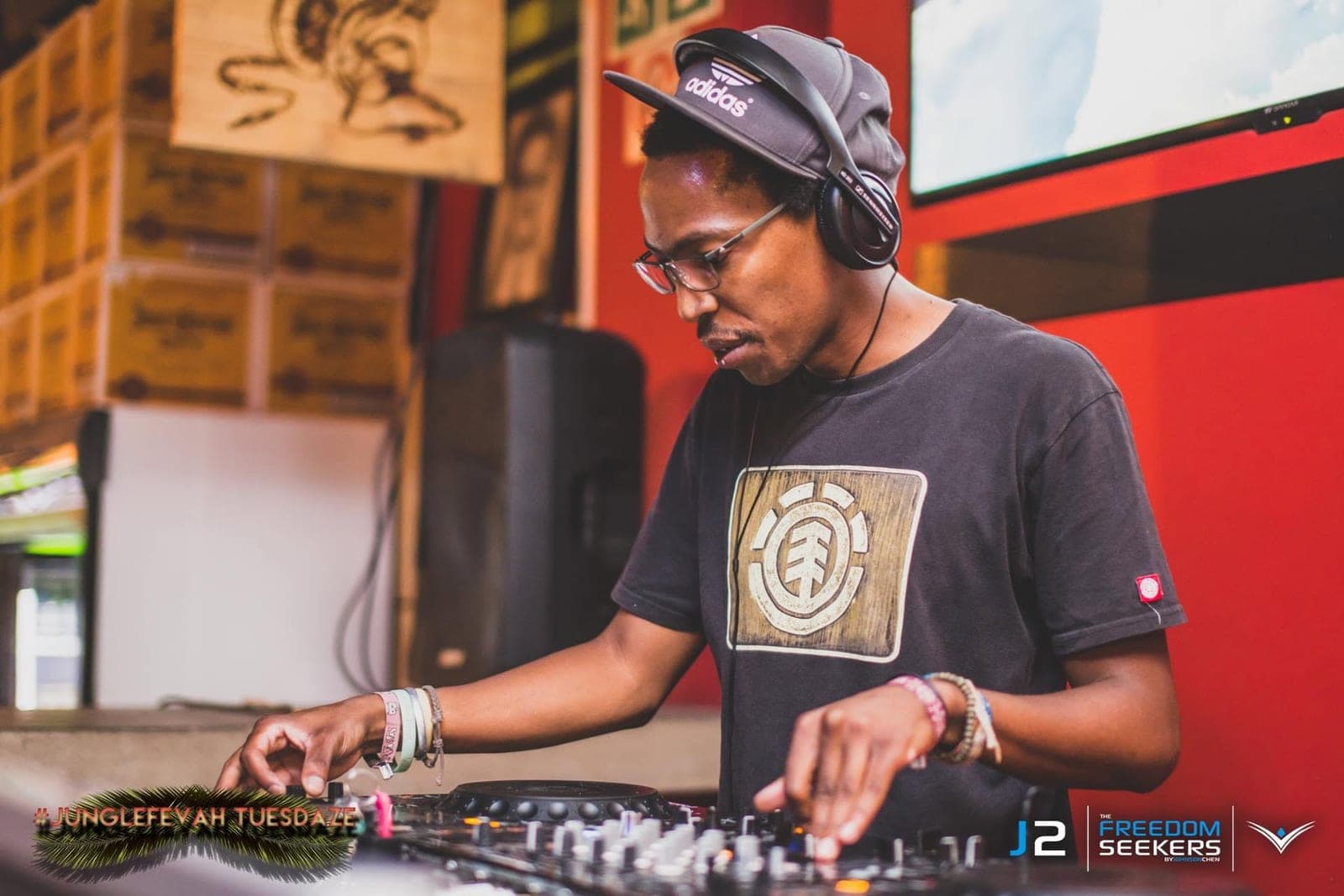 DJ in an adidas cap, glasses and headphones mixing at a bar with a red wall behind.