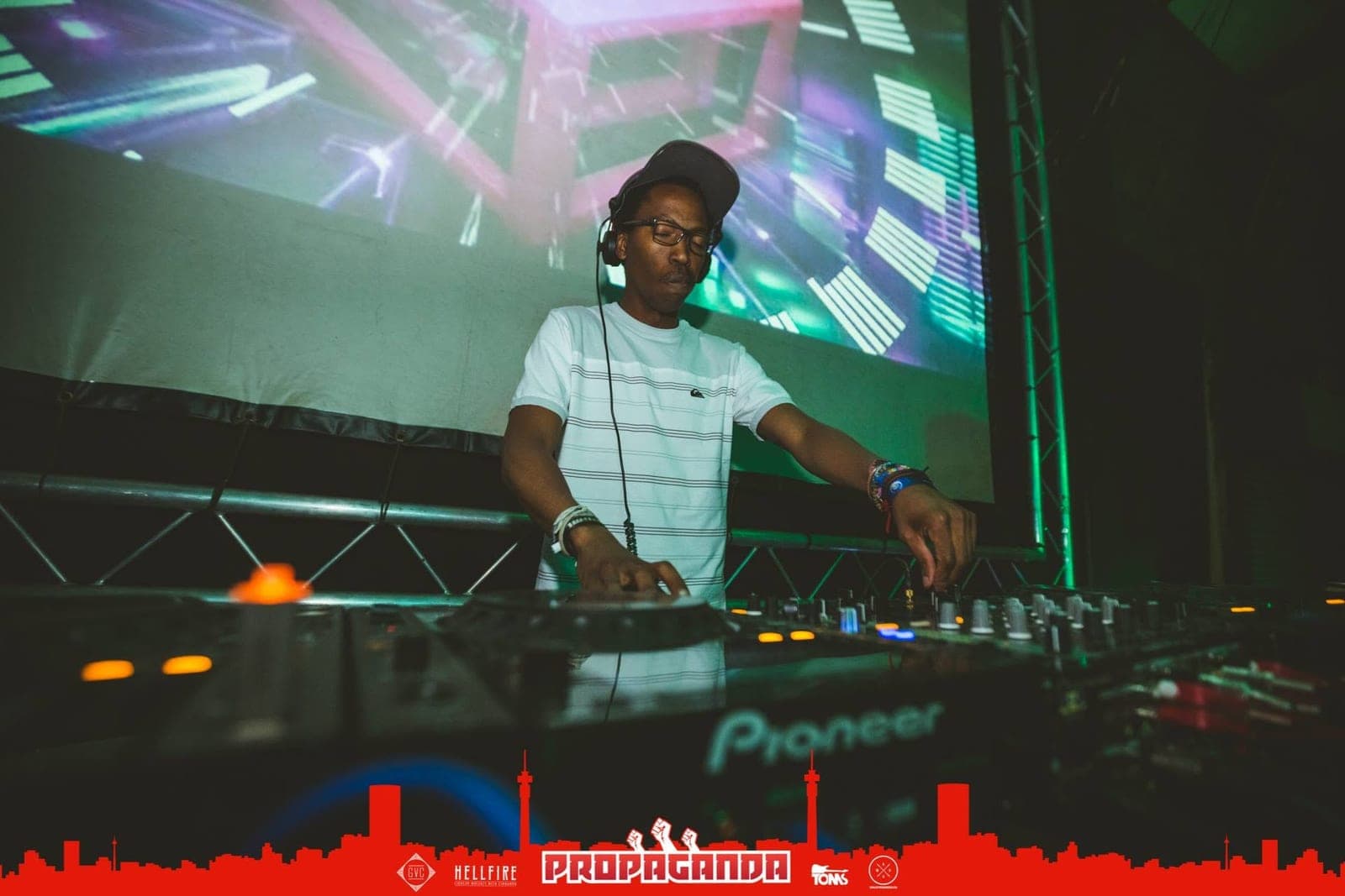 A DJ in a white striped t-shirt and cap, eyes closed, mixing in front of a large video screen.