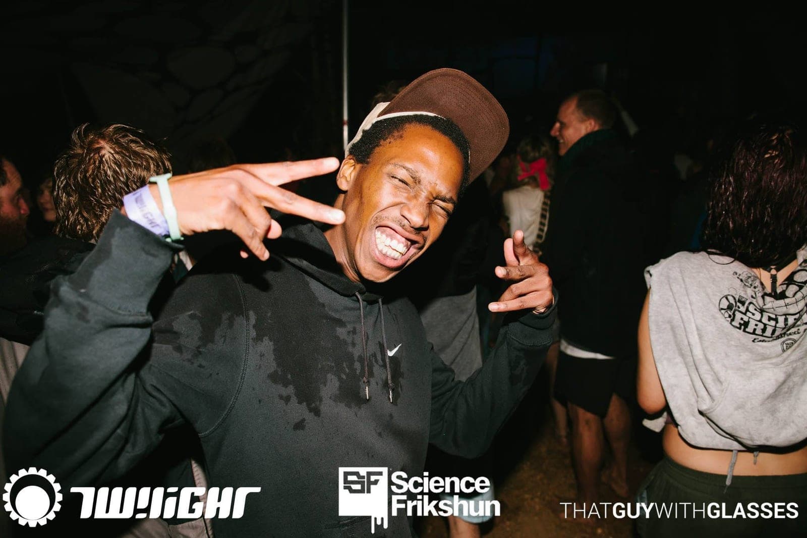 A grinning man in a brown cap flashing peace signs on a dark, crowded festival dance floor.